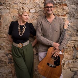 Un uomo e una donna con chitarre accanto a un muro di pietra.